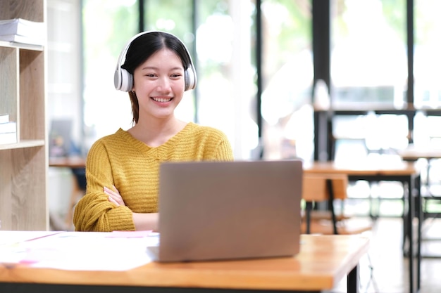 Young adult happy smiling Hispanic Asian student wearing headphones talking on online chat meeting using laptop in university campus or at virtual office College female student learning remotely