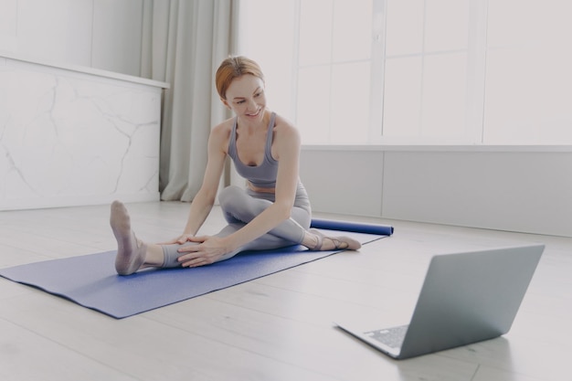 Young adult happy woman enjoying yoga practising stretching asana by online lesson