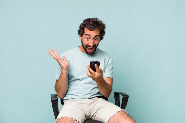 Young adult hispanic crazy man using his phone sitting on a chair