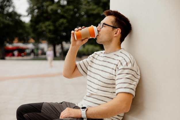 Young adult man sitting outdoors drinking coffee enjoying good weather and city view Man has a break from work spending time outside and relaxing Time with yourself dreaming mental health