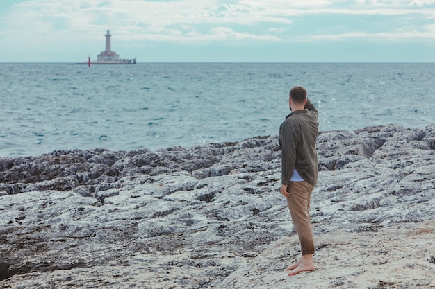 Young adult man walking by rocky sea beach barefoot summer vacation