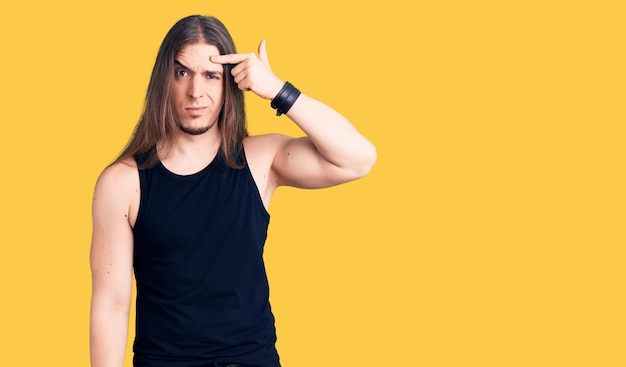 Young adult man with long hair wearing goth style with black clothes pointing unhappy to pimple on forehead ugly infection of blackhead acne and skin problem