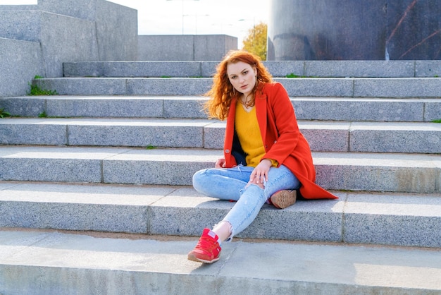 Young adult redhead woman in red coat blue jeans and red sneakers sits