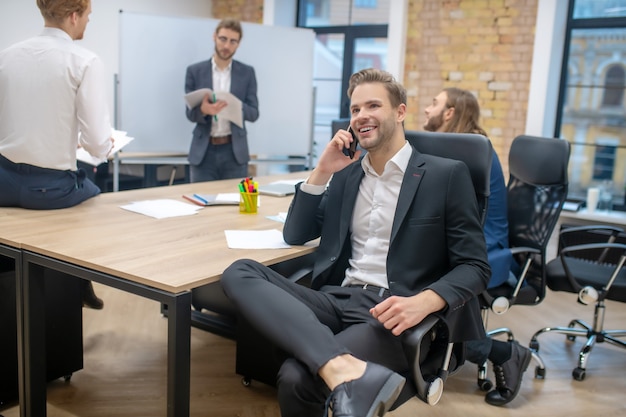 Young adult smiling man with smartphone in armchair and colleagues chatting after presentation