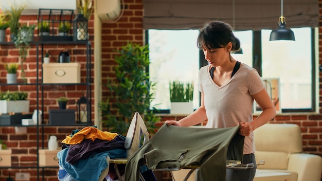 Young adult using steam iron to smooth out wrinkles on clothing, ironing pile of laundered clothes on ironing table. Casual person doing spring cleaning at home, housekeeping work.