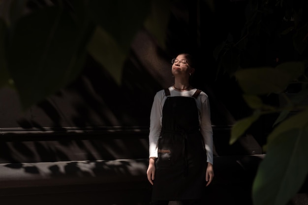 young adult woman in apron stands outdoors in shade of foliage