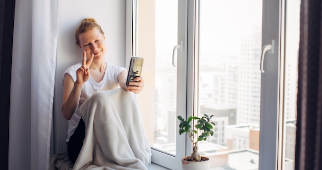 Young adult woman with glasses takes a selfie sitting on the windowsill at home, gesturing with her hand. Female smiles while chatting, looking at smartphone.