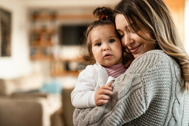 Young affectionate mother embracing her small daughter while spending time with her at home