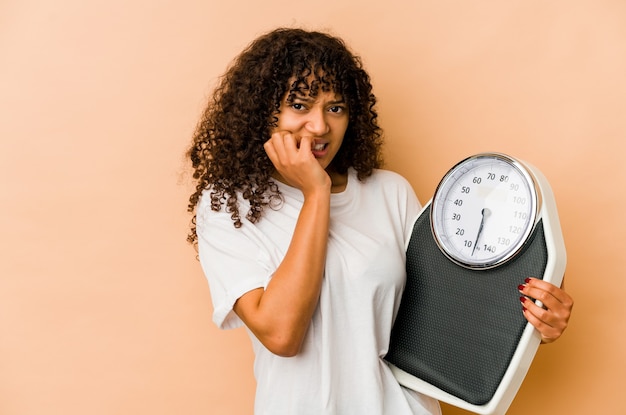 Young african american afro woman holding a scale biting fingernails, nervous and very anxious.