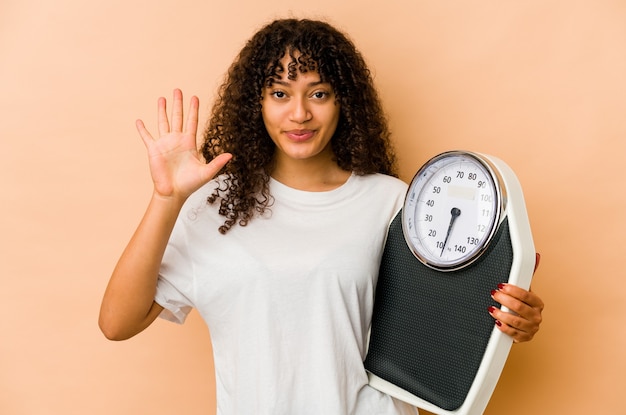 Young african american afro woman holding a scale smiling cheerful showing number five with fingers.