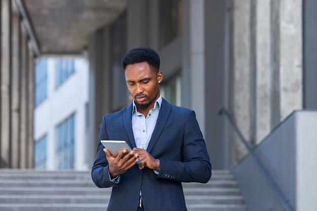 Young african american businessman in formal business suit standing working with tablet in hands on background modern office building outside. Man using smartphone or mobile phone outdoors city street