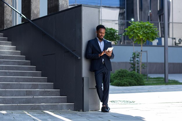 Young african american businessman in formal business suit standing working with tablet in hands on background modern office building outside. Man using smartphone or mobile phone outdoors city street