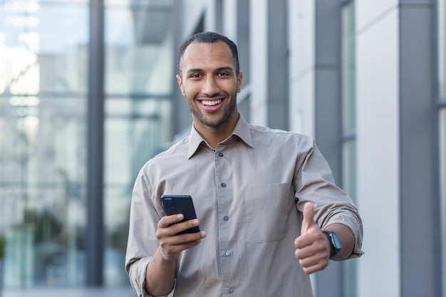 Young african american businessman outside office building smiling and looking at camera thumbs up