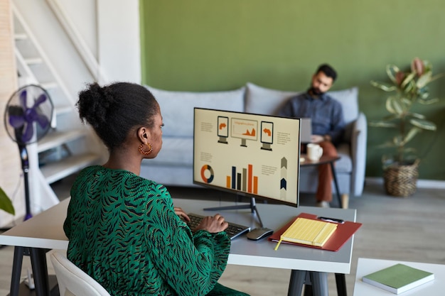 Young african american designer sitting in front of computer screen with graphic data and typing on