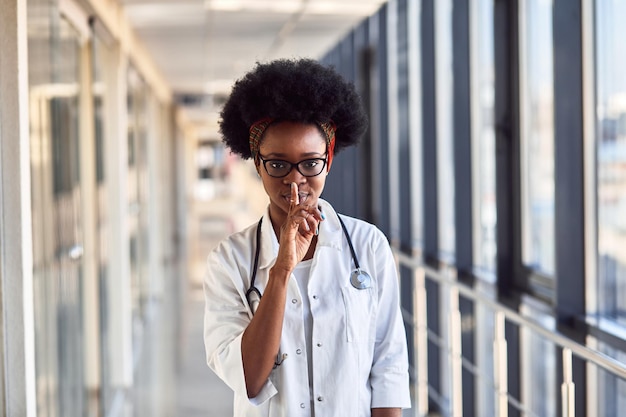 Young african american female doctor in white uniform standing in corridor and showing silence gesture.