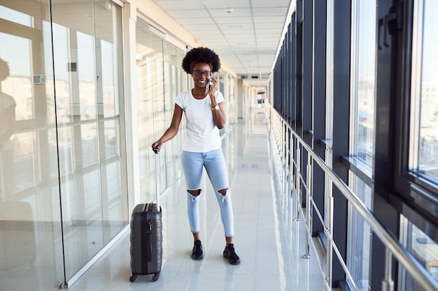 Young african american female passanger in casual clothes is in airport with baggage. Using phone.