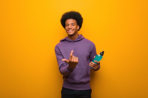 Young african american fitness man holding an energy drink inviting to come