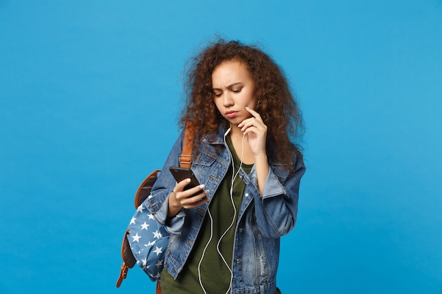 Young african american girl teen student in denim clothes, backpack headphones isolated on blue wall