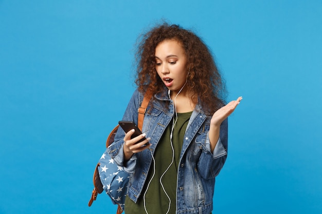 Young african american girl teen student in denim clothes, backpack headphones isolated on blue wall