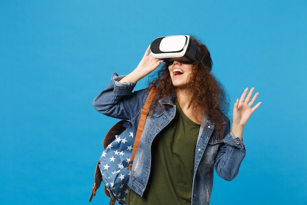 Young african american girl teen student in denim clothes, backpack headset vr isolated on blue wall