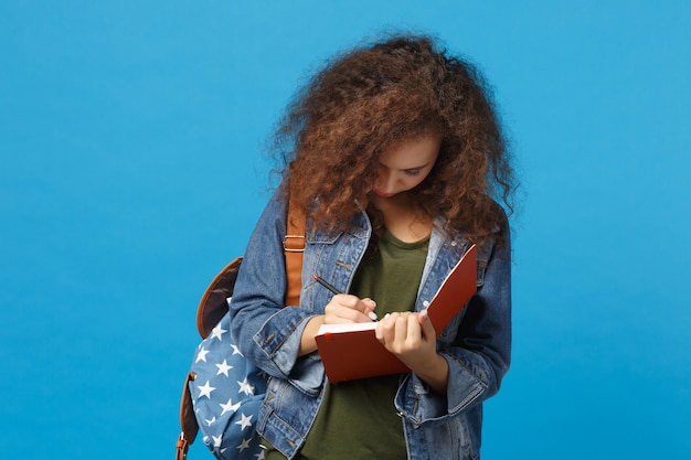 Young african american girl teen student in denim clothes, backpack hold books isolated on blue wall