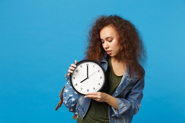 Young african american girl teen student in denim clothes, backpack hold clock isolated on blue wall