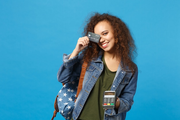 Young african american girl teen student in denim clothes, backpack hold credit card isolated on blue wall