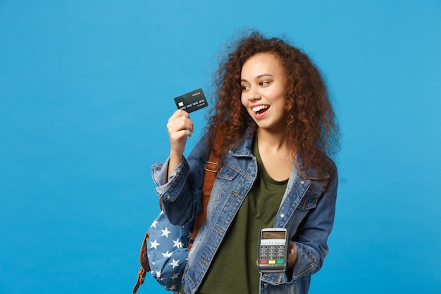 Young african american girl teen student in denim clothes, backpack hold credit card isolated on blue wall