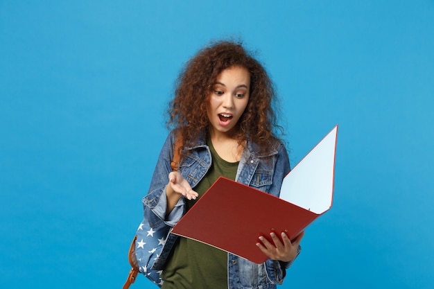 Young african american girl teen student in denim clothes backpack hold folder isolated on blue wall