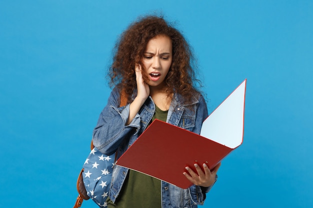 Young african american girl teen student in denim clothes backpack hold folder isolated on blue wall