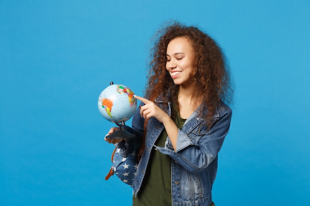 Young african american girl teen student in denim clothes, backpack hold globe isolated on blue wall