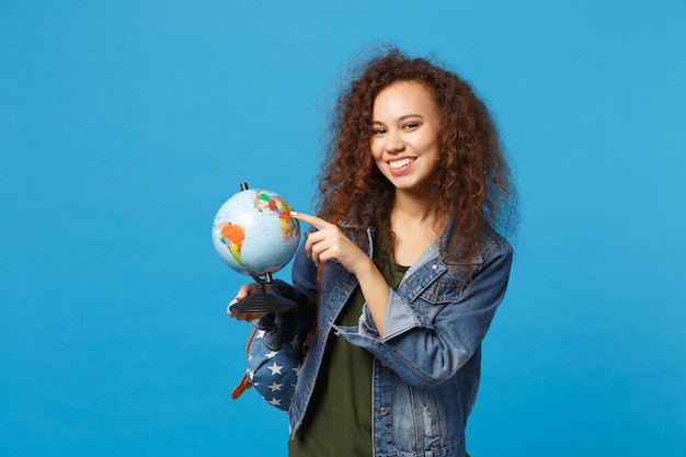 Young african american girl teen student in denim clothes, backpack hold globe isolated on blue wall