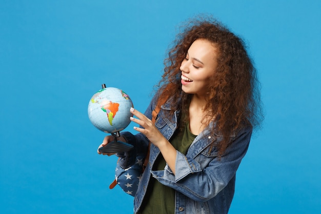 Young african american girl teen student in denim clothes, backpack hold globe isolated on blue wall