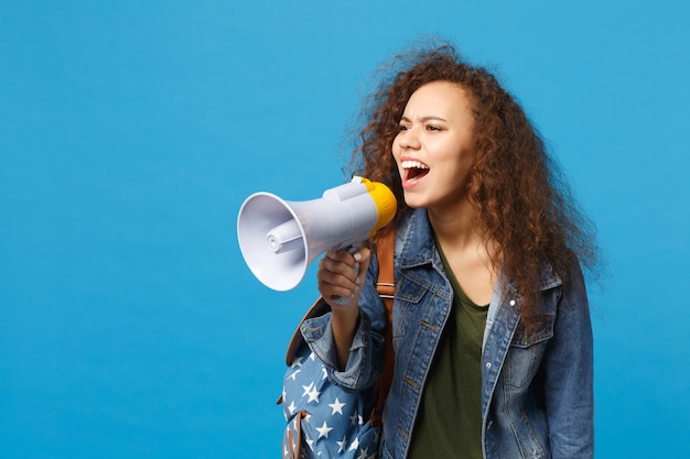 Young african american girl teen student in denim clothes, backpack hold megaphone isolated on blue wall