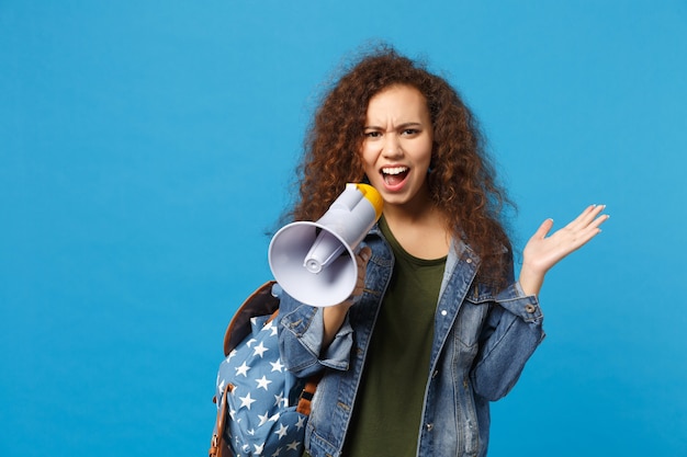 Young african american girl teen student in denim clothes, backpack hold megaphone isolated on blue wall