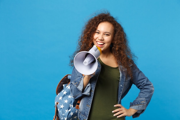 Young african american girl teen student in denim clothes, backpack hold megaphone isolated on blue wall