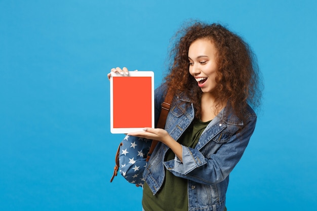 Young african american girl teen student in denim clothes, backpack hold pad pc isolated on blue wall wall