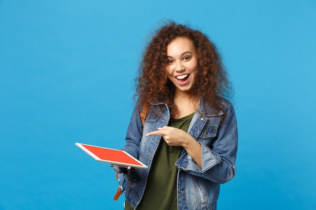 Young african american girl teen student in denim clothes, backpack hold pad pc isolated on blue wall wall