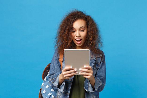 Young african american girl teen student in denim clothes, backpack hold pad pc isolated on blue wall wall