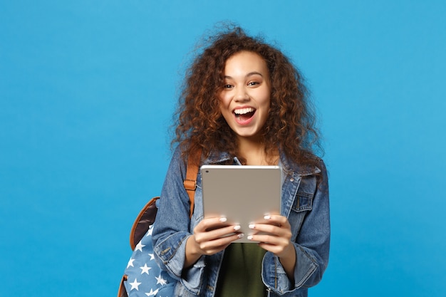 Young african american girl teen student in denim clothes, backpack hold pad pc isolated on blue wall wall