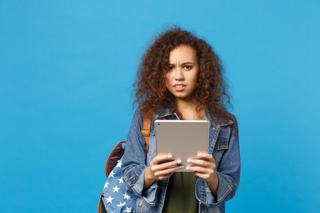 Young african american girl teen student in denim clothes, backpack hold pad pc isolated on blue wall wall