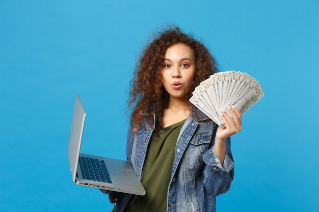 Young african american girl teen student in denim clothes, backpack hold pc, fan of cash money isolated on blue wall