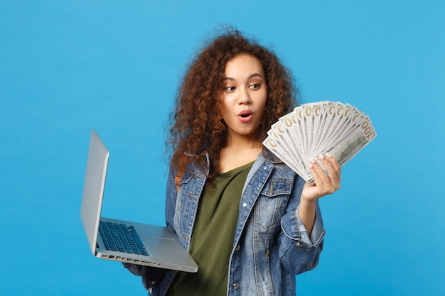 Young african american girl teen student in denim clothes, backpack hold pc, fan of cash money isolated on blue wall