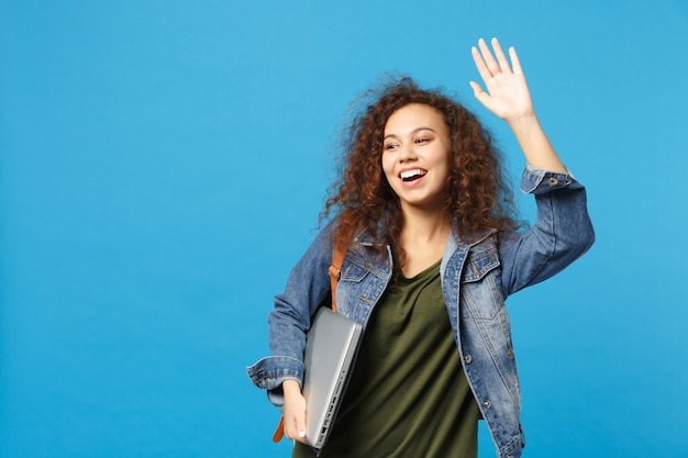 Young african american girl teen student in denim clothes, backpack hold pc isolated on blue wall