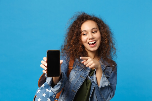 Young african american girl teen student in denim clothes, backpack hold phone isolated on blue wall