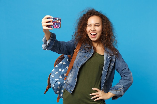 Young african american girl teen student in denim clothes, backpack hold phone isolated on blue wall