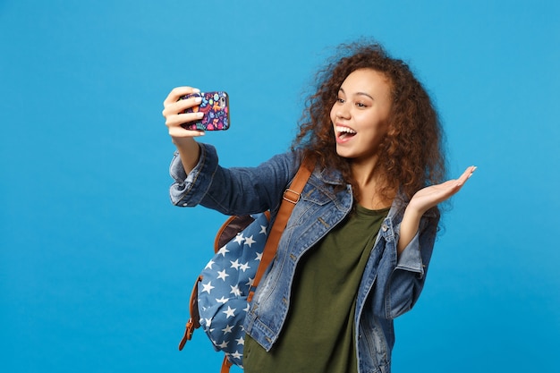 Young african american girl teen student in denim clothes, backpack hold phone isolated on blue wall