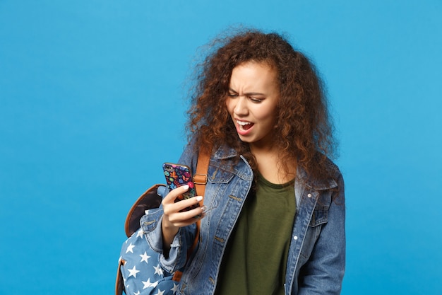 Young african american girl teen student in denim clothes, backpack hold phone isolated on blue wall