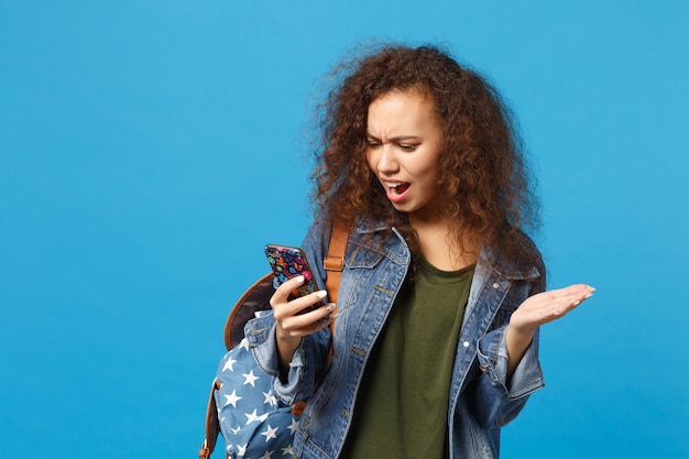 Young african american girl teen student in denim clothes, backpack hold phone isolated on blue wall