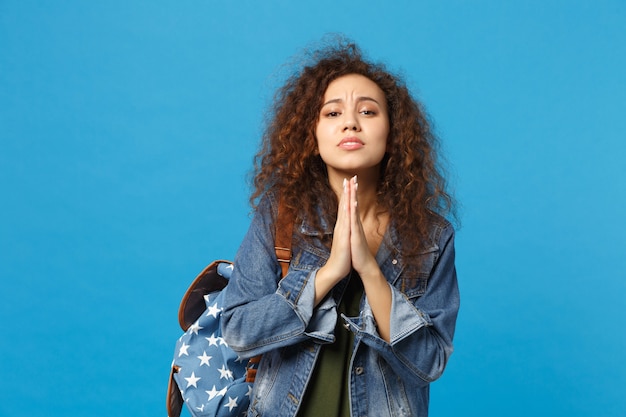 Young african american girl teen student in denim clothes, backpack making wish isolated on blue wall